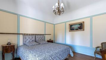 Une chambre à coucher comprenant un lit, un lustre, une table de chevet, une lampe, un tableau encadré et une chaise.