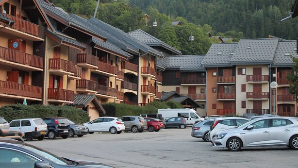 A multi-story hotel with balconies, a parking lot with various cars, and a mountainous backdrop. - Studio for 4, balcony, shared parking (Valloire)