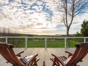 A wooden deck with two lounge chairs facing a grassy field and a tree, under a partly cloudy sky. - The Schooner (Goleen)