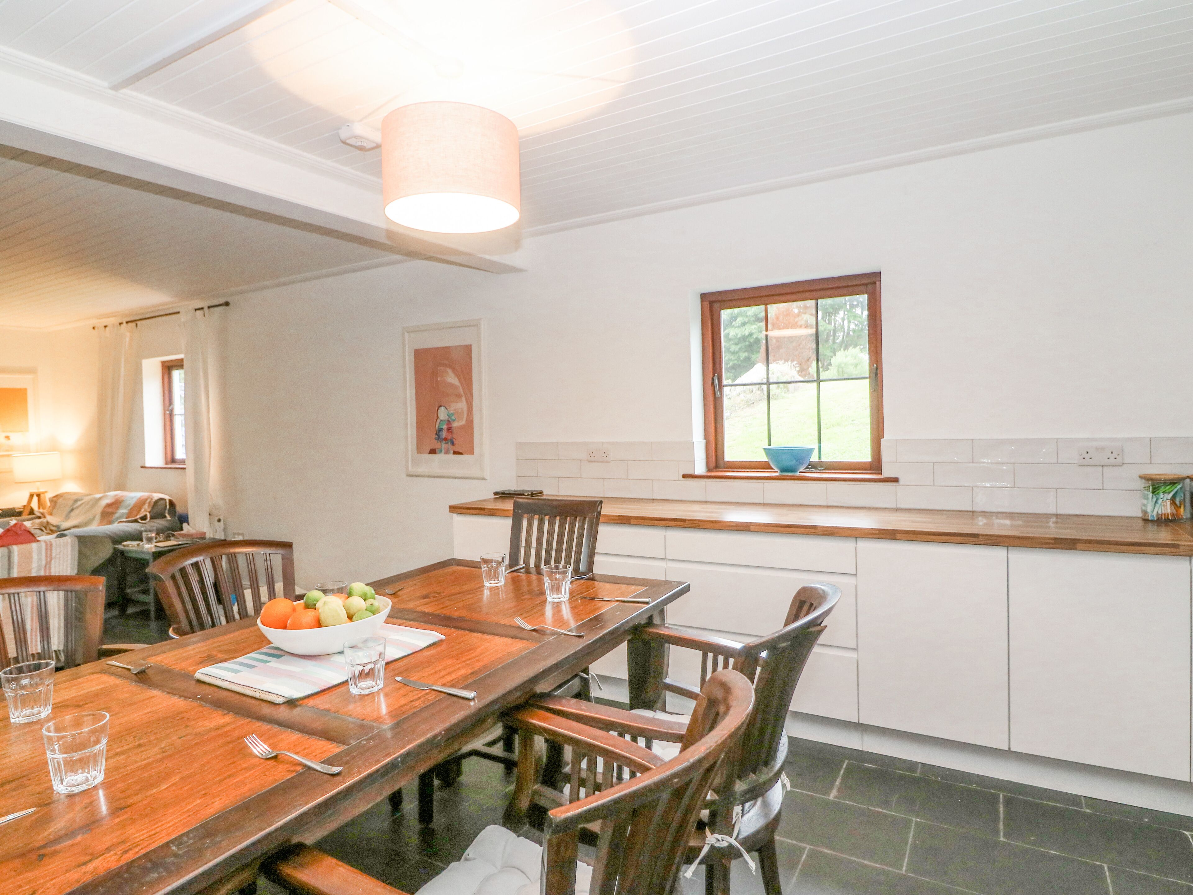 A dining area with a wooden table, chairs, and a bowl of fruit. There is a window with a view of greenery outside.