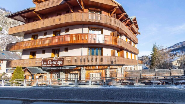 En flerlags bygning i træ med balkoner, et skilt der viser 'La Grange Restaurant La Grange', og et bjerglandskab i baggrunden.