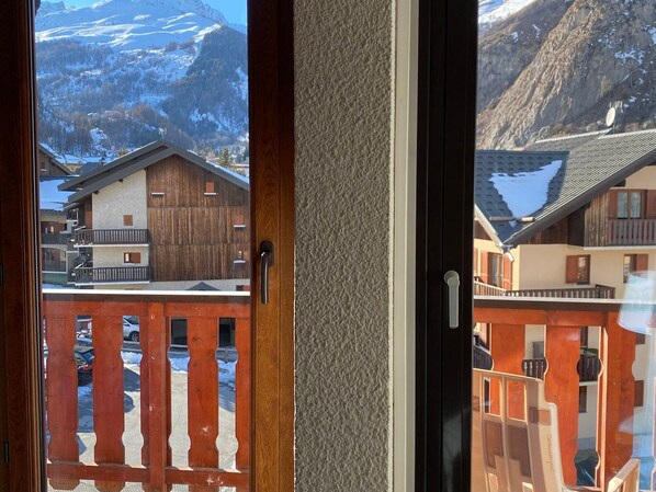 A view through a door with a wooden frame and a glass panel, revealing a mountain landscape with snow-covered peaks and buildings with wooden facades and balconies.