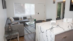 A modern living room with a grey sofa, a glass coffee table, and a marble kitchen island.