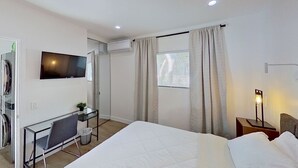 A hotel room with a bed, a desk with a chair, a television, an air conditioning unit, a window with curtains, and a bedside table with a lamp.