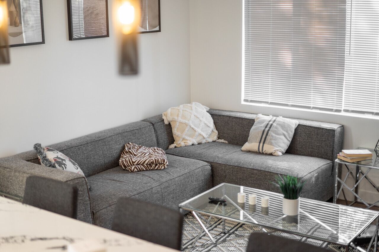 A modern living room with a grey sectional sofa, a glass coffee table, and a window with blinds.