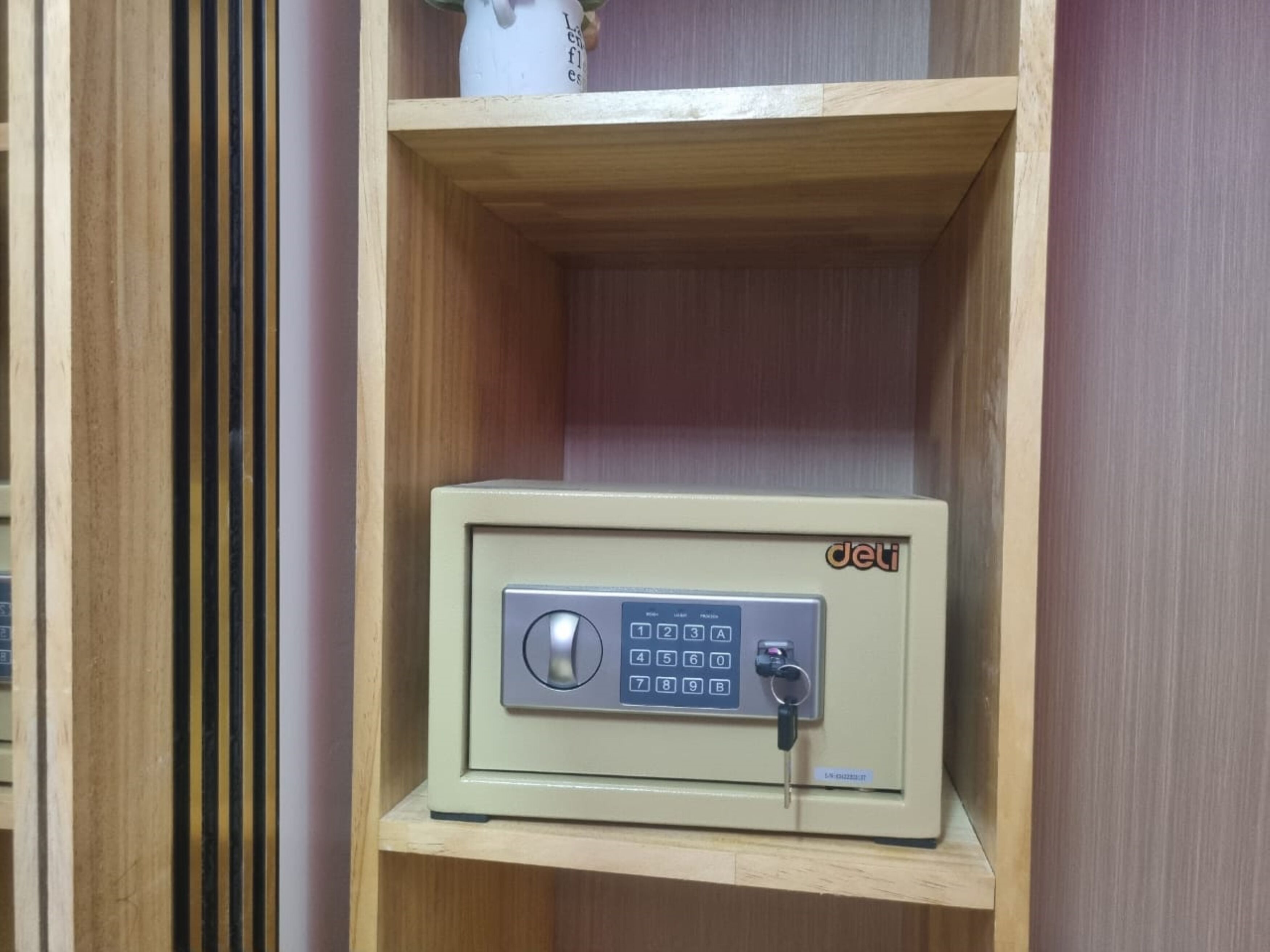 Ein beiger Safe mit Tastatur und Schlüsselring auf einem Holzregal in einem Raum mit vertikalen Rollläden.