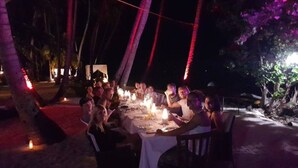 A group of people dining at an outdoor table with candles, surrounded by palm trees and a beach setting. - Batuta Maldives Inn (Thulusdhoo Island)