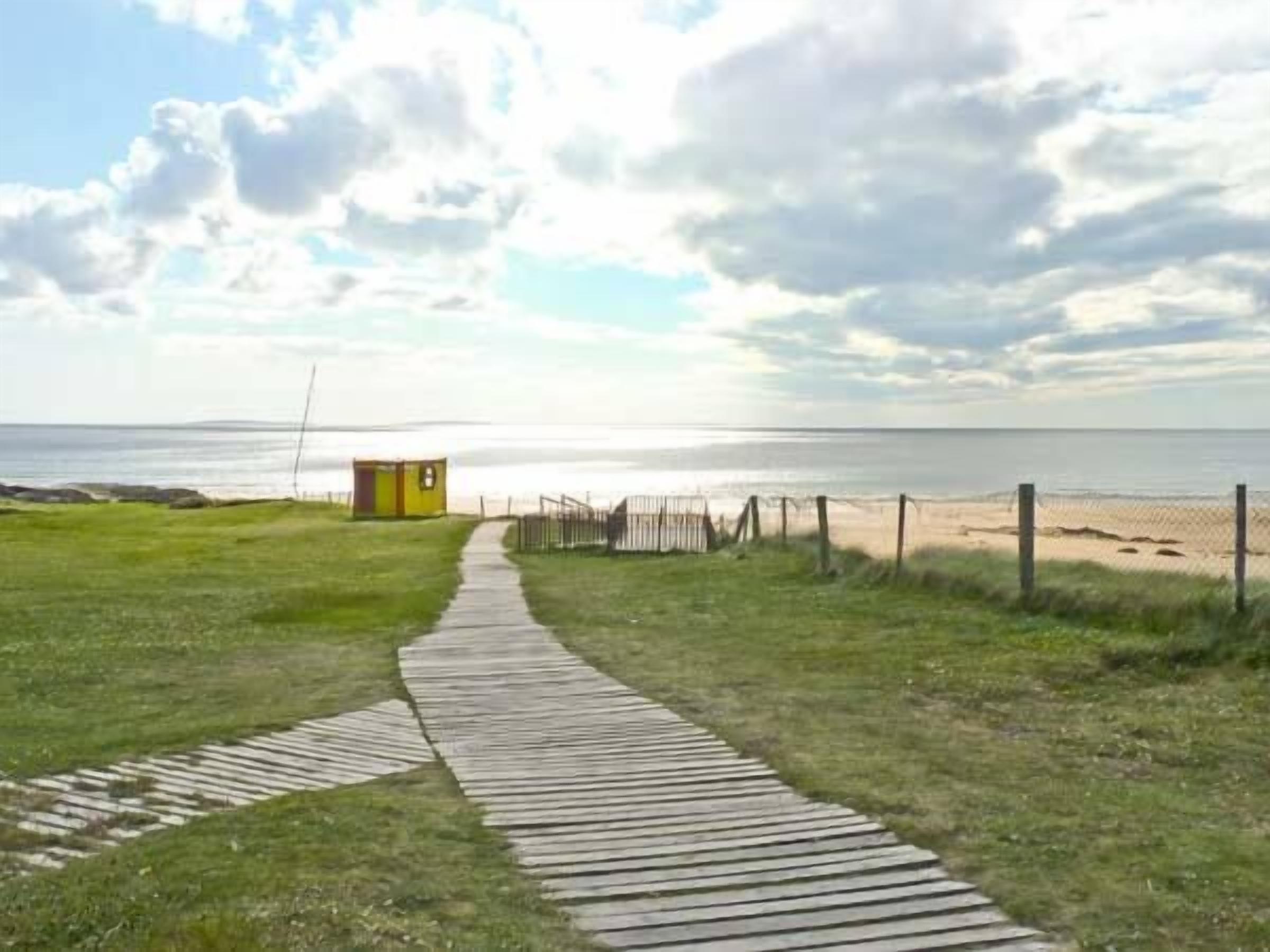 A wooden pathway leading to a beach with a small yellow structure, likely a restroom, and a fence along the shore.