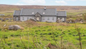 A stone house with a grey roof, set in a grassy field with rolling hills in the background. - Cúnna Bán (Fanore)