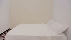 Ein kleines, ordentlich bezogenes Bett in einem Eckzimmer mit schlichten Wänden und einem gemusterten Boden.