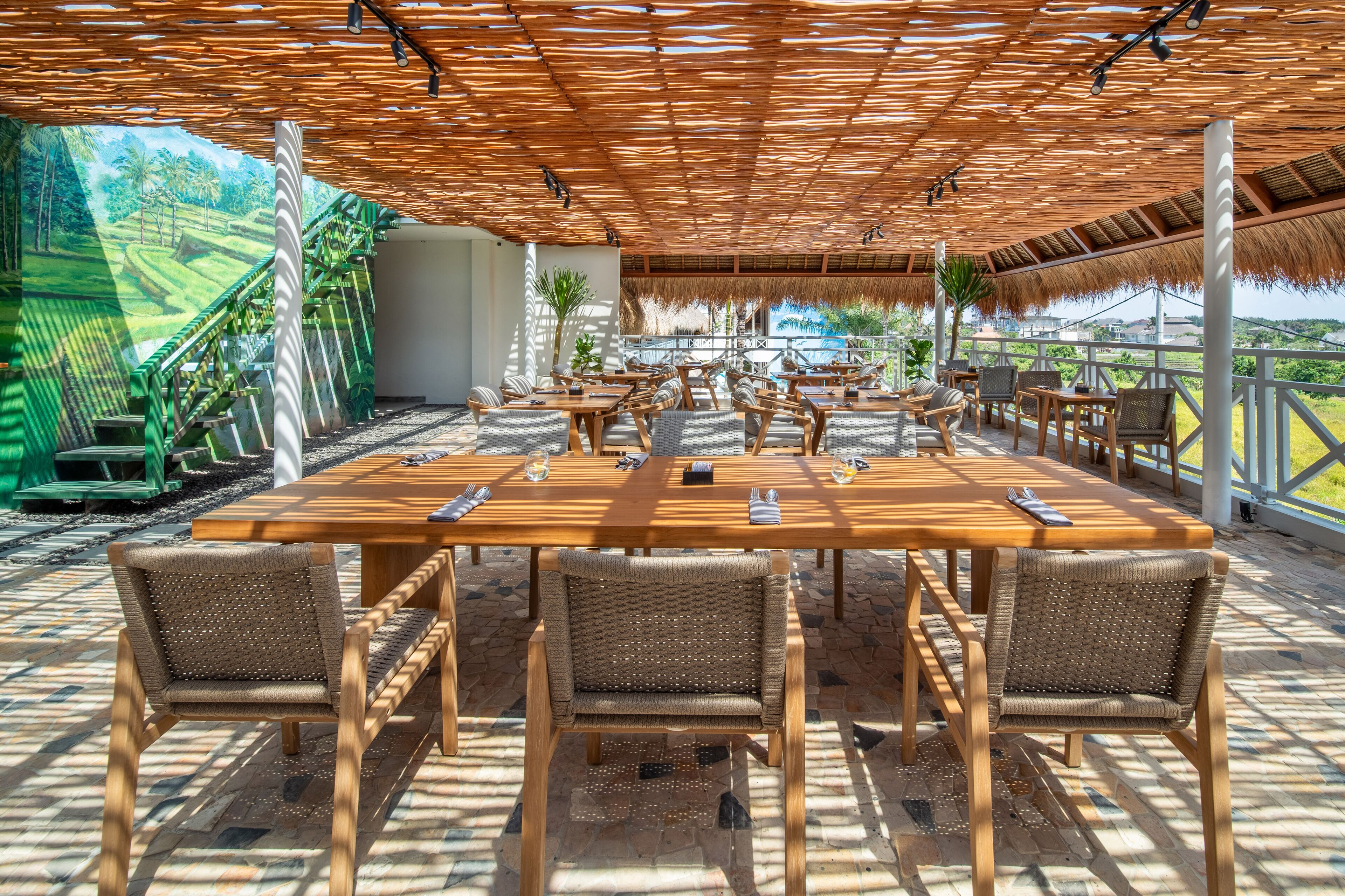 An outdoor dining area with a long wooden table and wicker chairs, surrounded by a thatched roof and a scenic view.