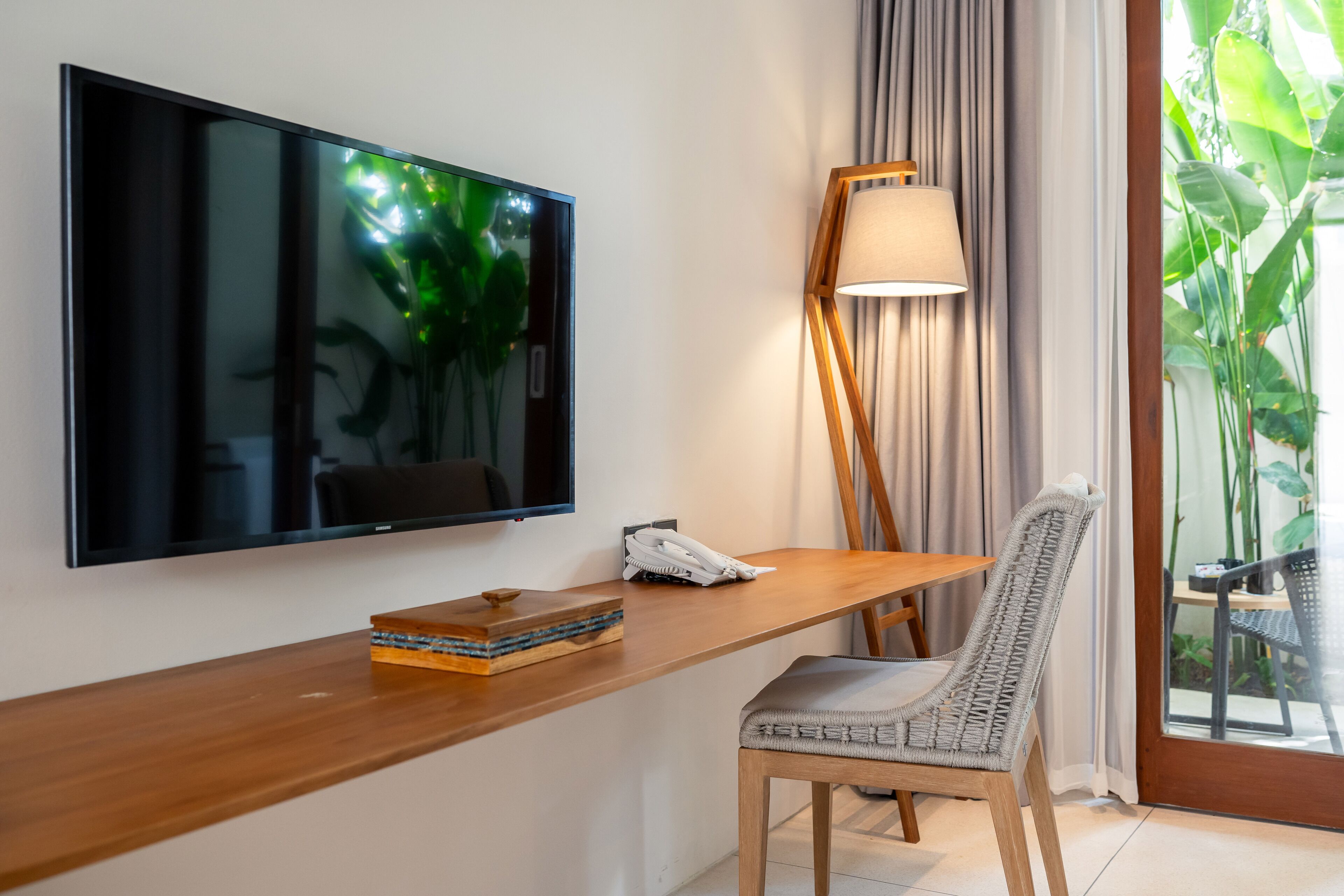 Una habitación moderna con una televisión de pantalla plana montada en la pared, un escritorio de madera con una lámpara, una silla y una mesita con un teléfono y un libro.