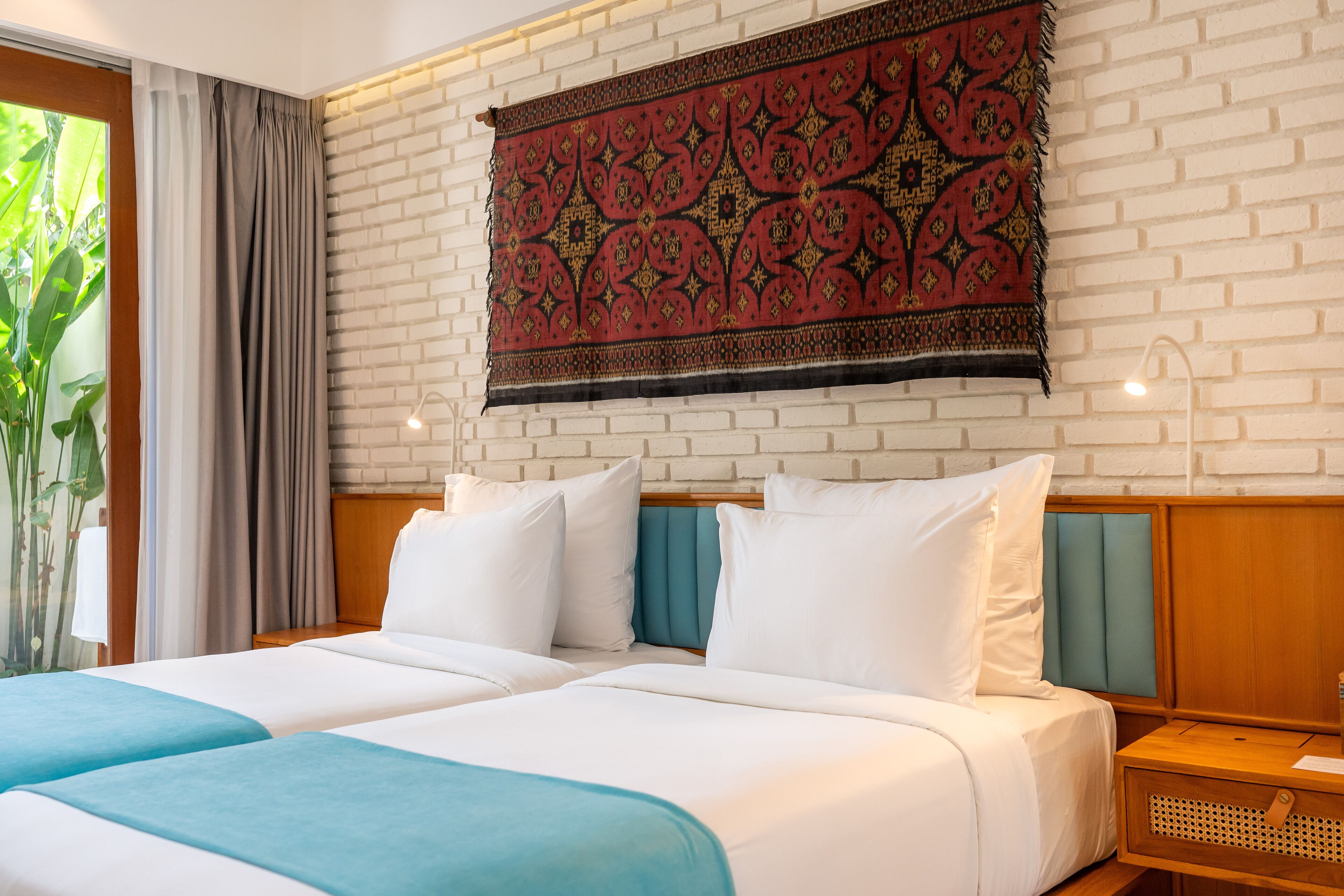 A double bed with white and teal bedding, a headboard with teal panels, and a red patterned tapestry on a brick wall.