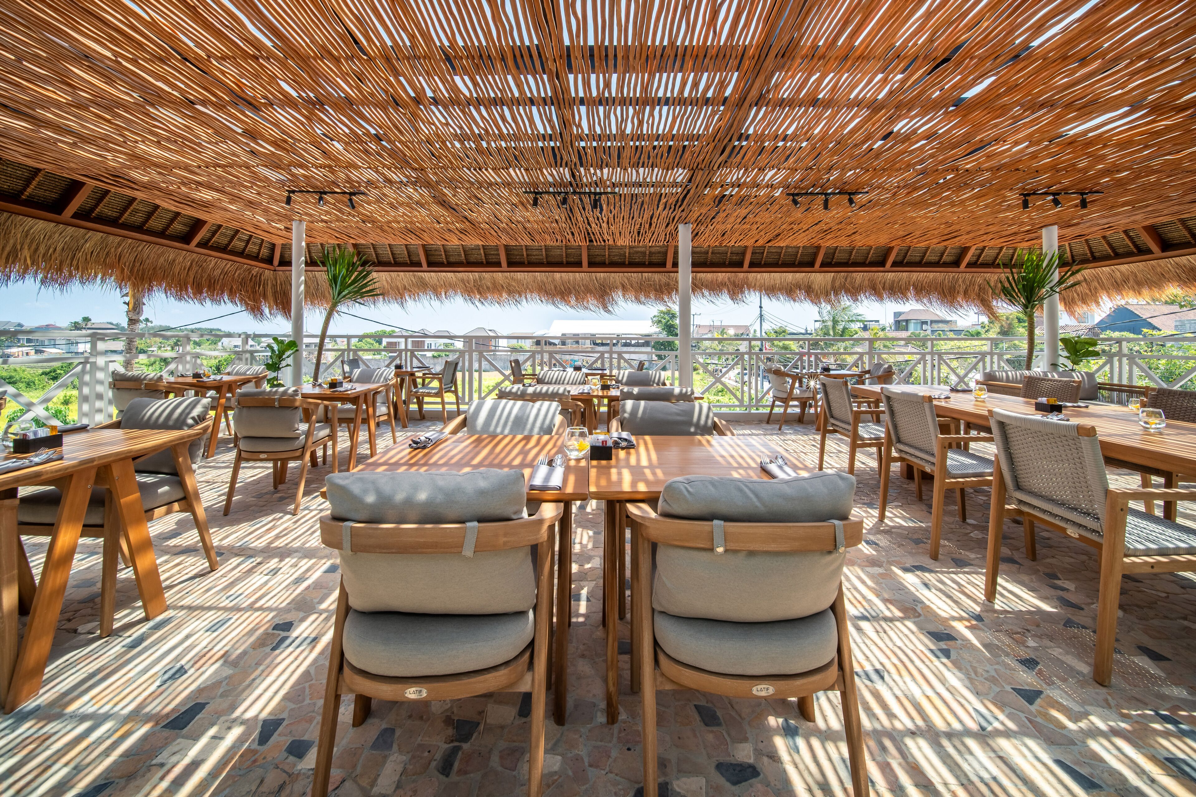An outdoor dining area with wooden tables and chairs, a thatched roof, and a view of the landscape.