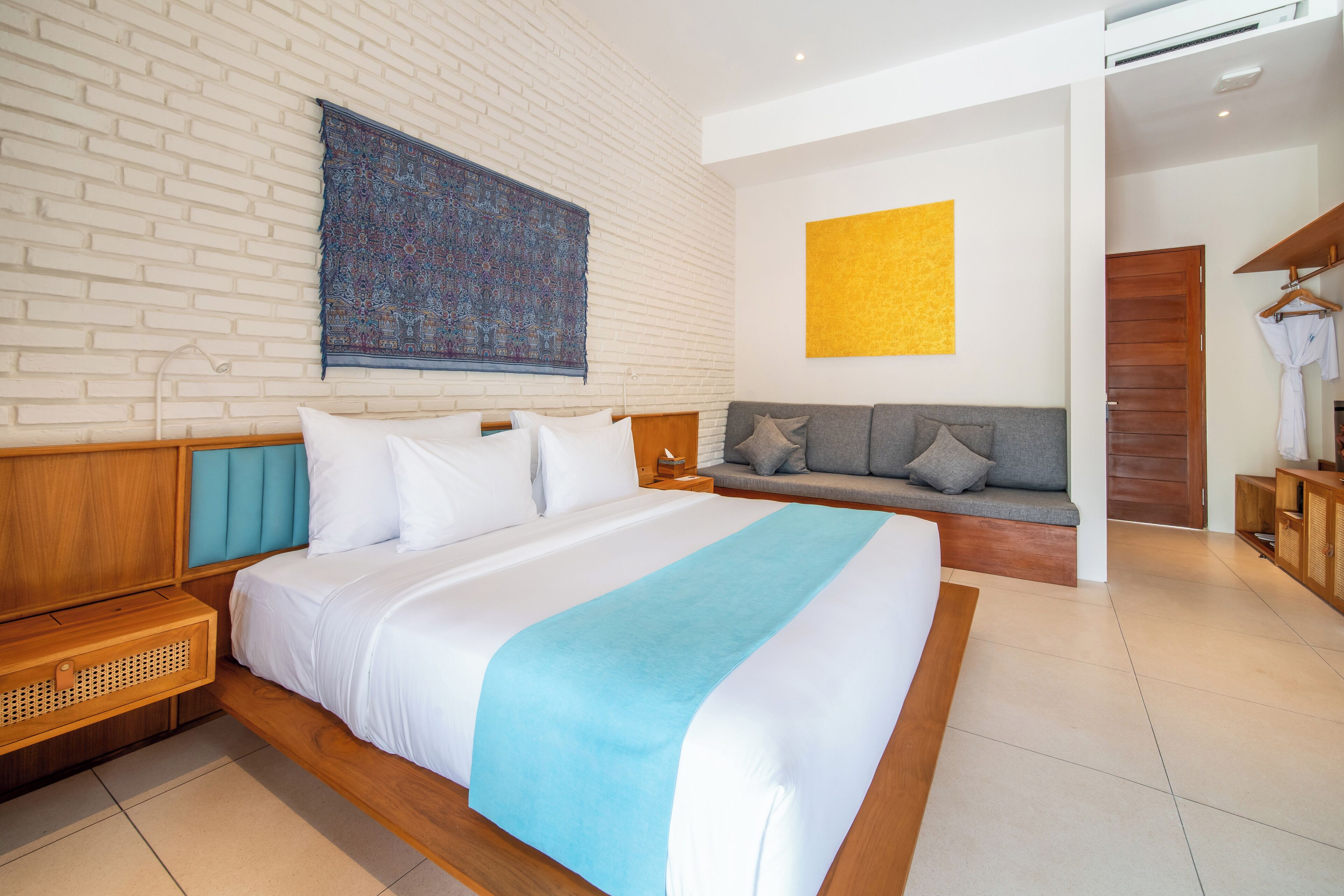 Una habitación de hotel moderna con una cama grande, un sofá gris, un cabecero de madera, un tapiz estampado y un cuadro abstracto amarillo.