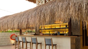 Uma área de bar com telhado de palha, banquetas de madeira e balcão com garrafas e copos.