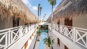 A hotel corridor with thatched-roof buildings, a central water feature, and a white railing.