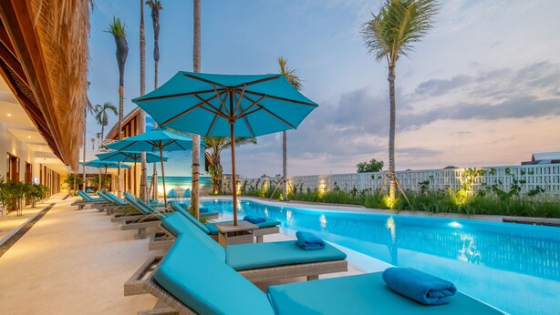Área da piscina com espreguiçadeiras e guarda-sóis, vista clara da piscina e um edifício com design moderno.