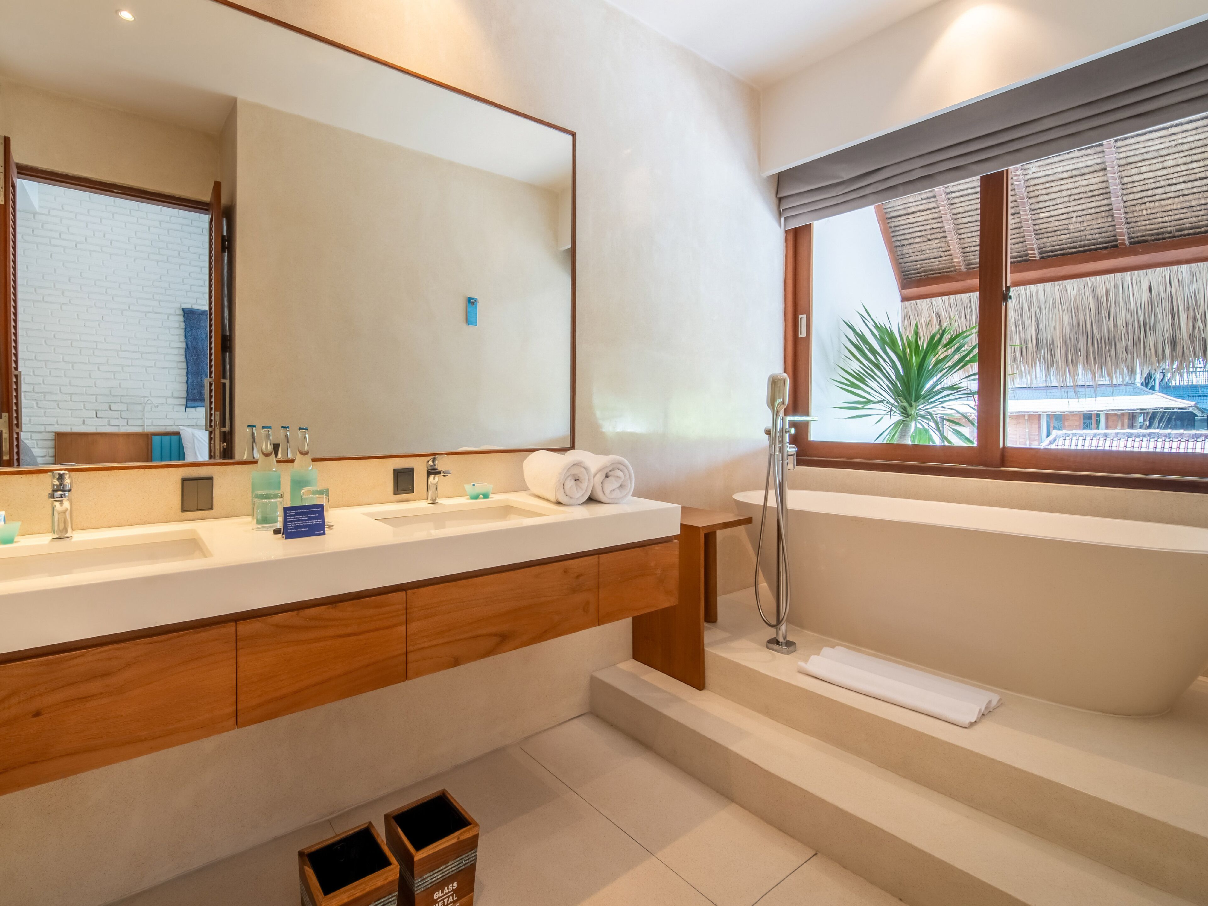 A modern bathroom with a large mirror, two sinks, a bathtub, and a window with a view of a building.