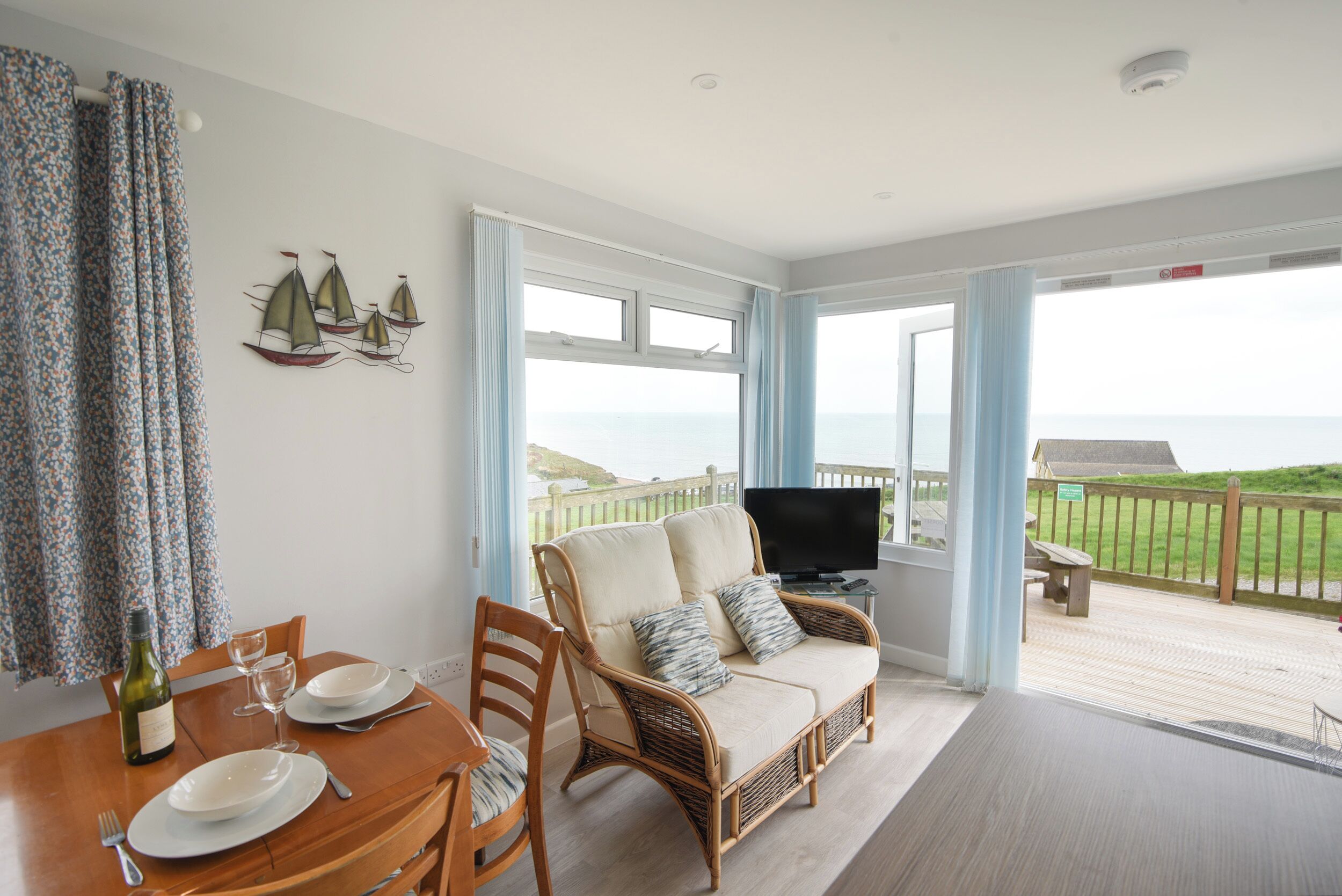 A dining area with a table set for two, a wicker sofa with cushions, a television on a stand, and a view of the sea through large windows.