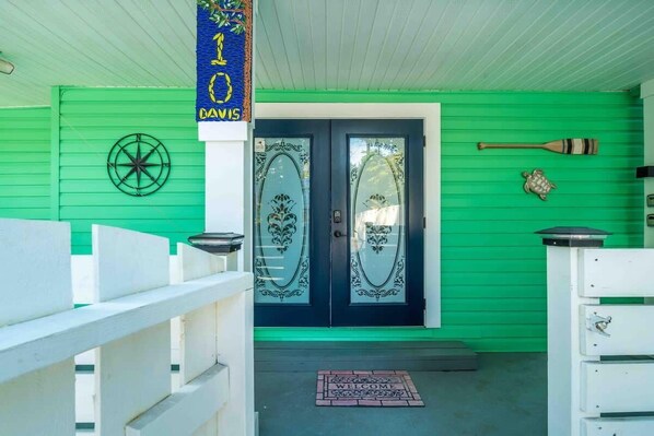 A green building with a white porch, a welcome mat, and a bat and turtle decoration on the wall.