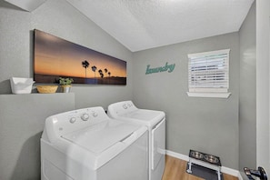 A laundry room with two white front-loading washers, a painting of a sunset, a wall-mounted laundry sign, and a pet feeding station.