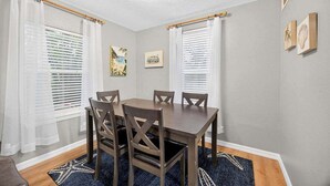 Uma sala de jantar com mesa e cadeiras de madeira, cortinas brancas e obras de arte nas paredes.