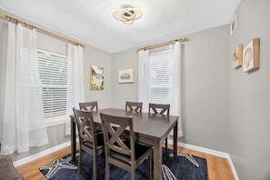 A dining room with a wooden table and chairs, white curtains, and artwork on the walls. - Magnolia Cove - Fire Pit, Hot Tub, Downtown (St. Augustine)