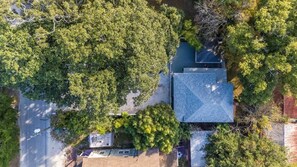 An aerial view of a residential area with a house featuring a sloped roof, surrounded by dense green trees.