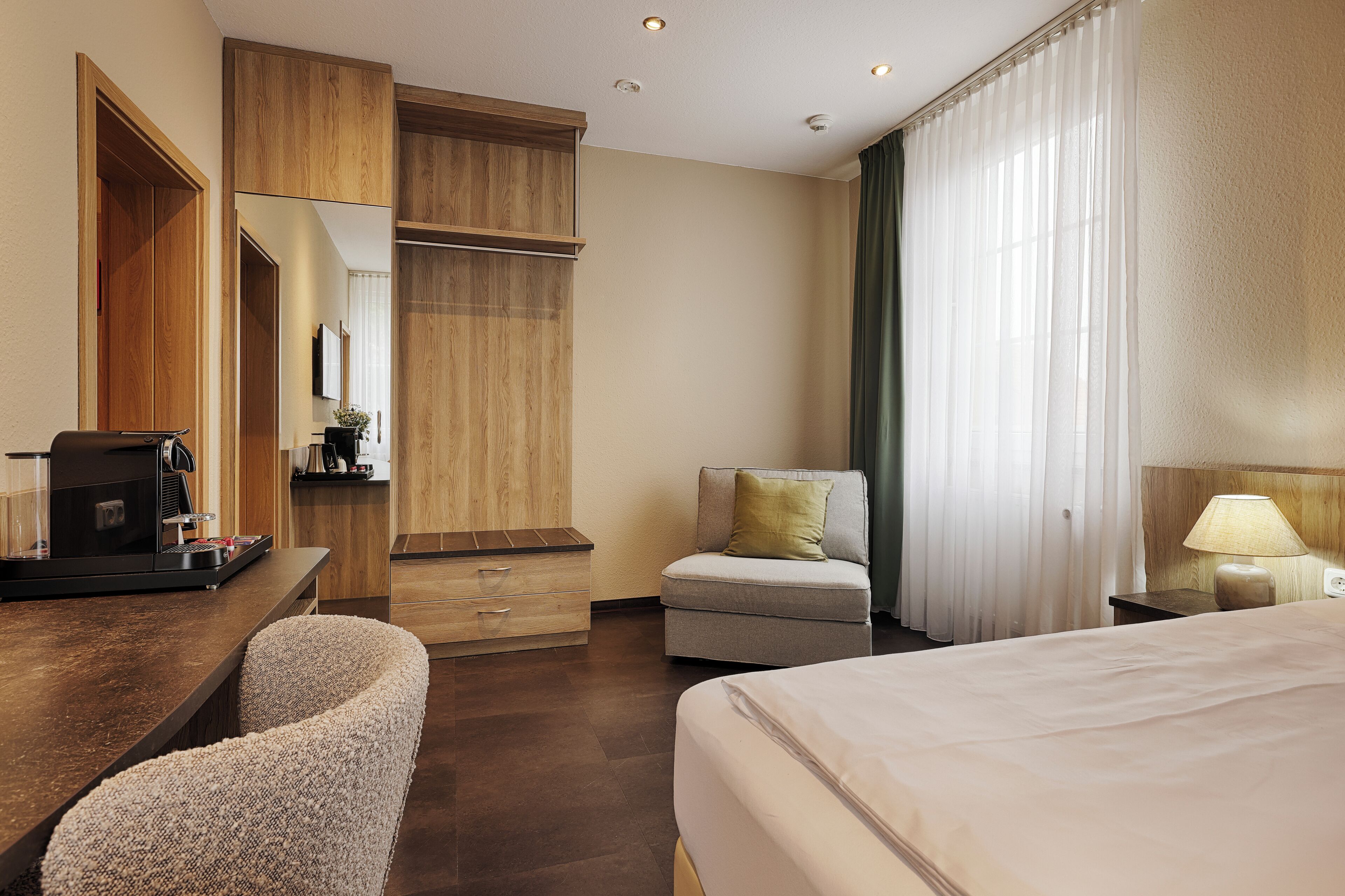 Habitación de hotel con cama, escritorio con cafetera, sillón, sofá y ventana grande con cortinas.