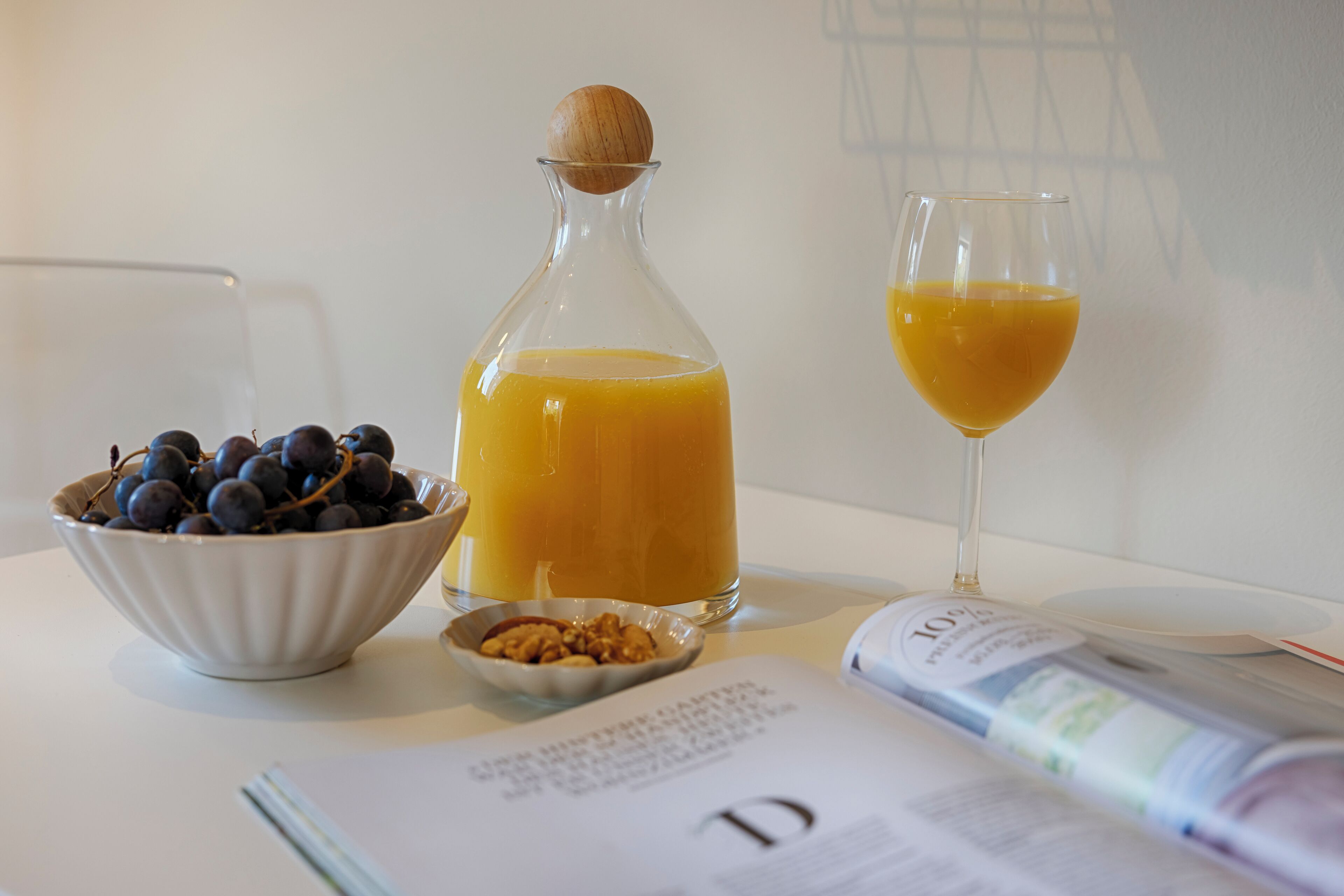 A table with a bowl of blueberries, a glass of orange juice, a bottle of juice, a small bowl of nuts, and a magazine.