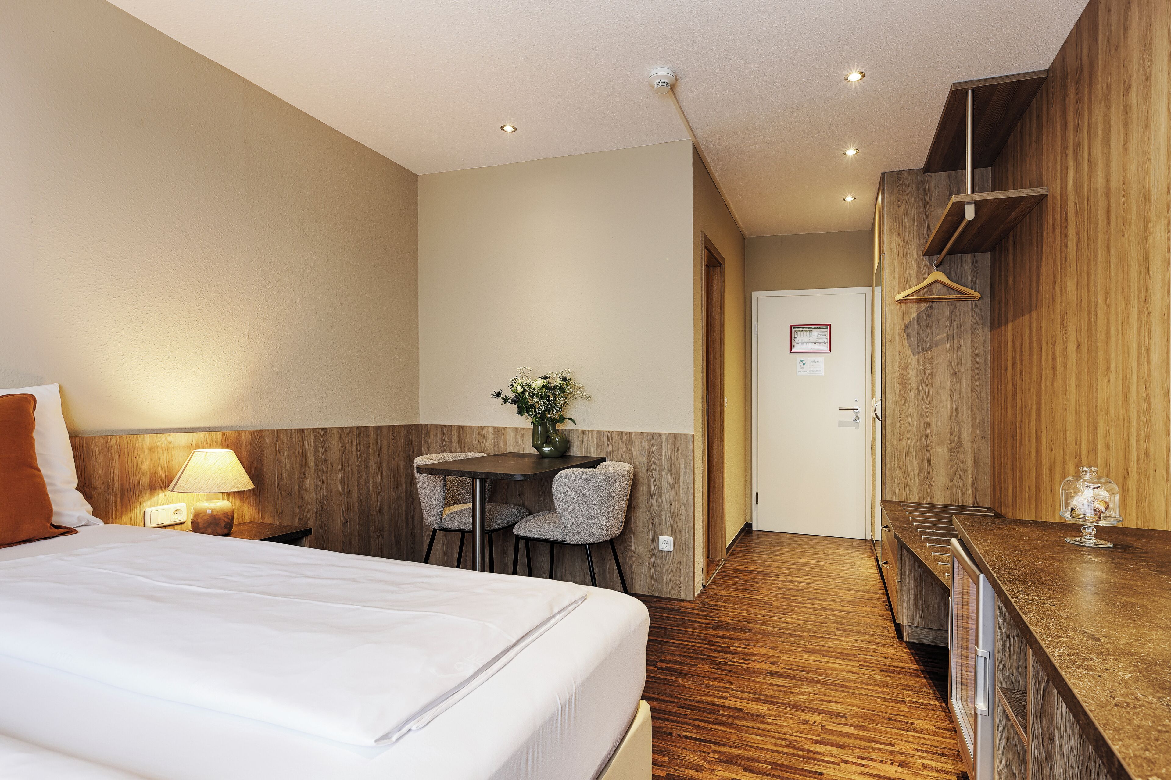 Habitación de hotel con cama, mesita de noche con lámpara, pequeña mesa de comedor con sillas, escritorio de madera y piso de madera.