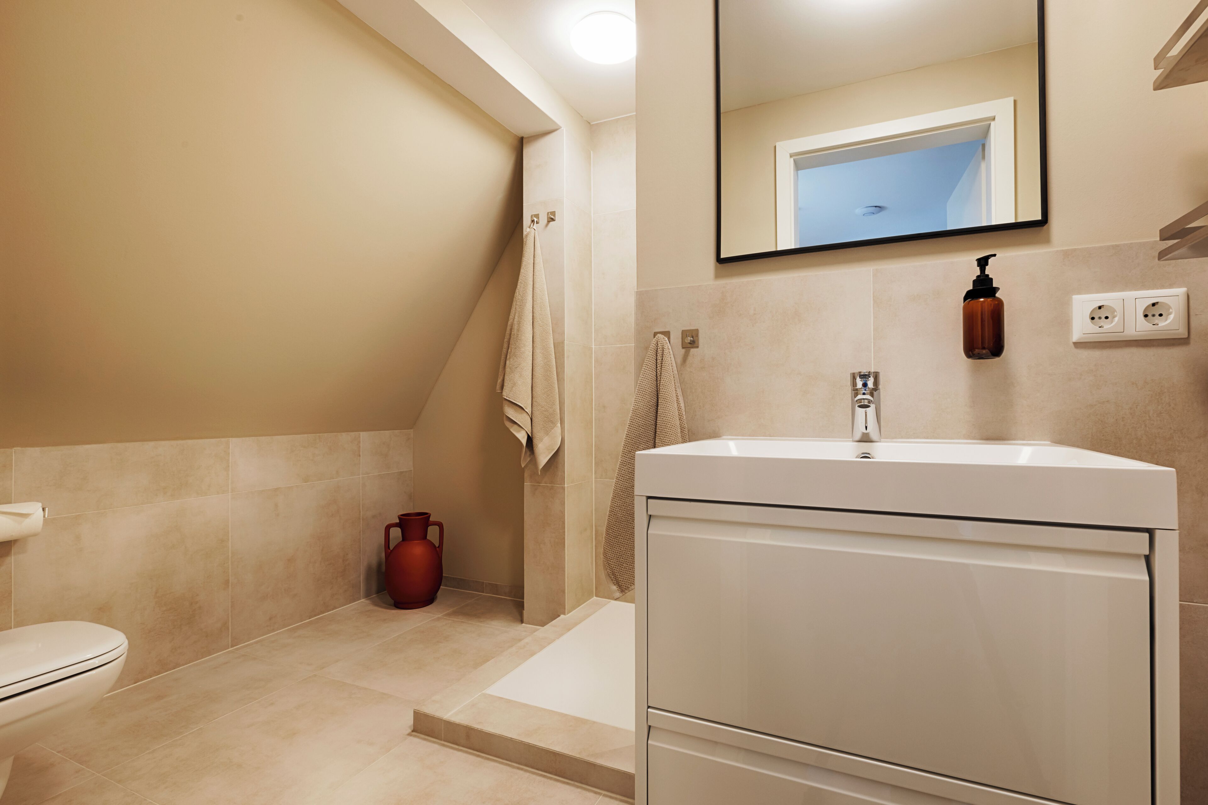Un baño moderno con un lavamanos blanco, un espejo grande y un jarrón rojo en el suelo.