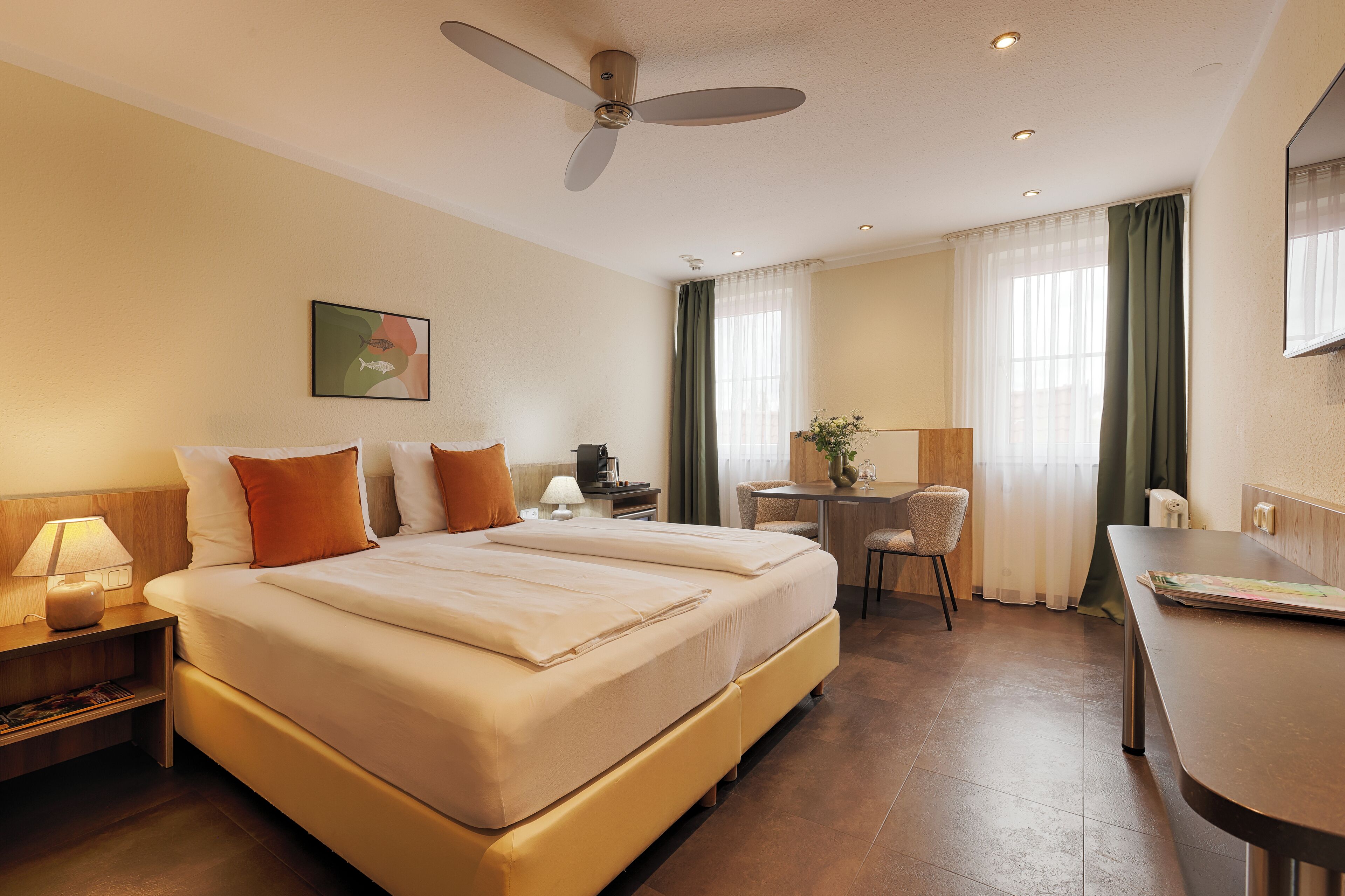 A hotel room with a large bed, a desk, a chair, a ceiling fan, and a painting on the wall.