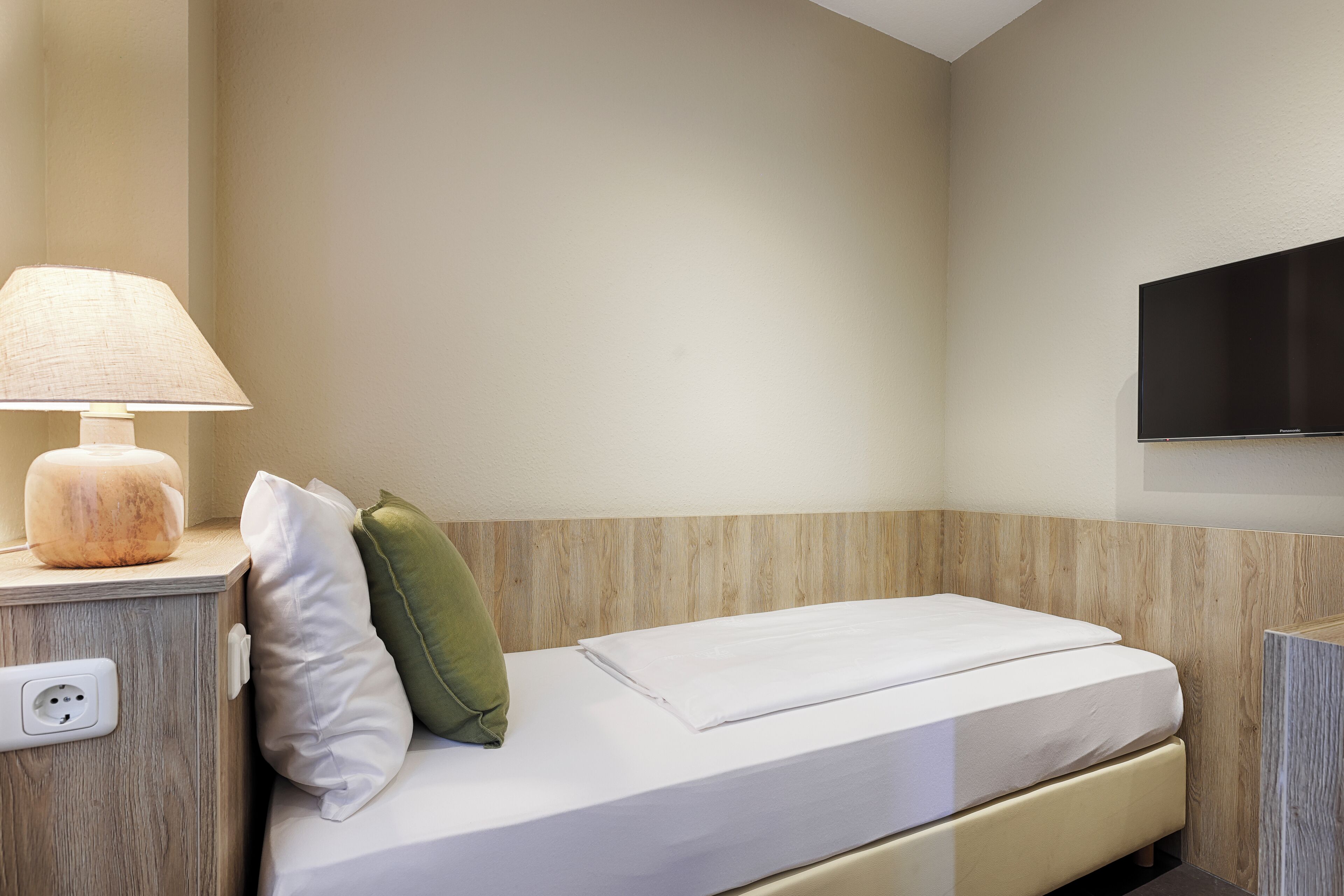 A neatly made bed with white bedding, a green pillow, and a lamp on a wooden nightstand.
