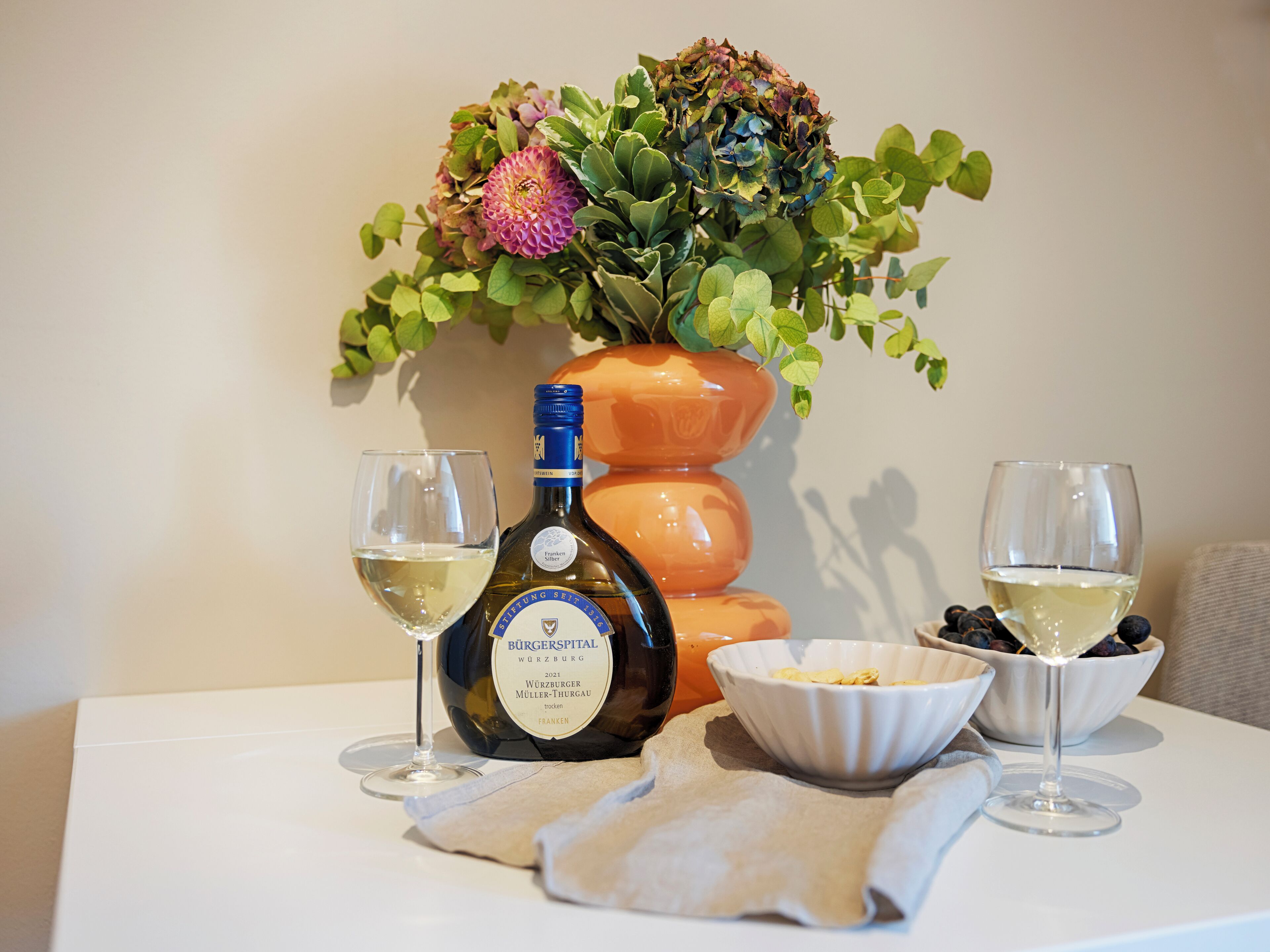 Una mesa con una botella de vino, dos copas, un bol de bayas y una planta decorativa.