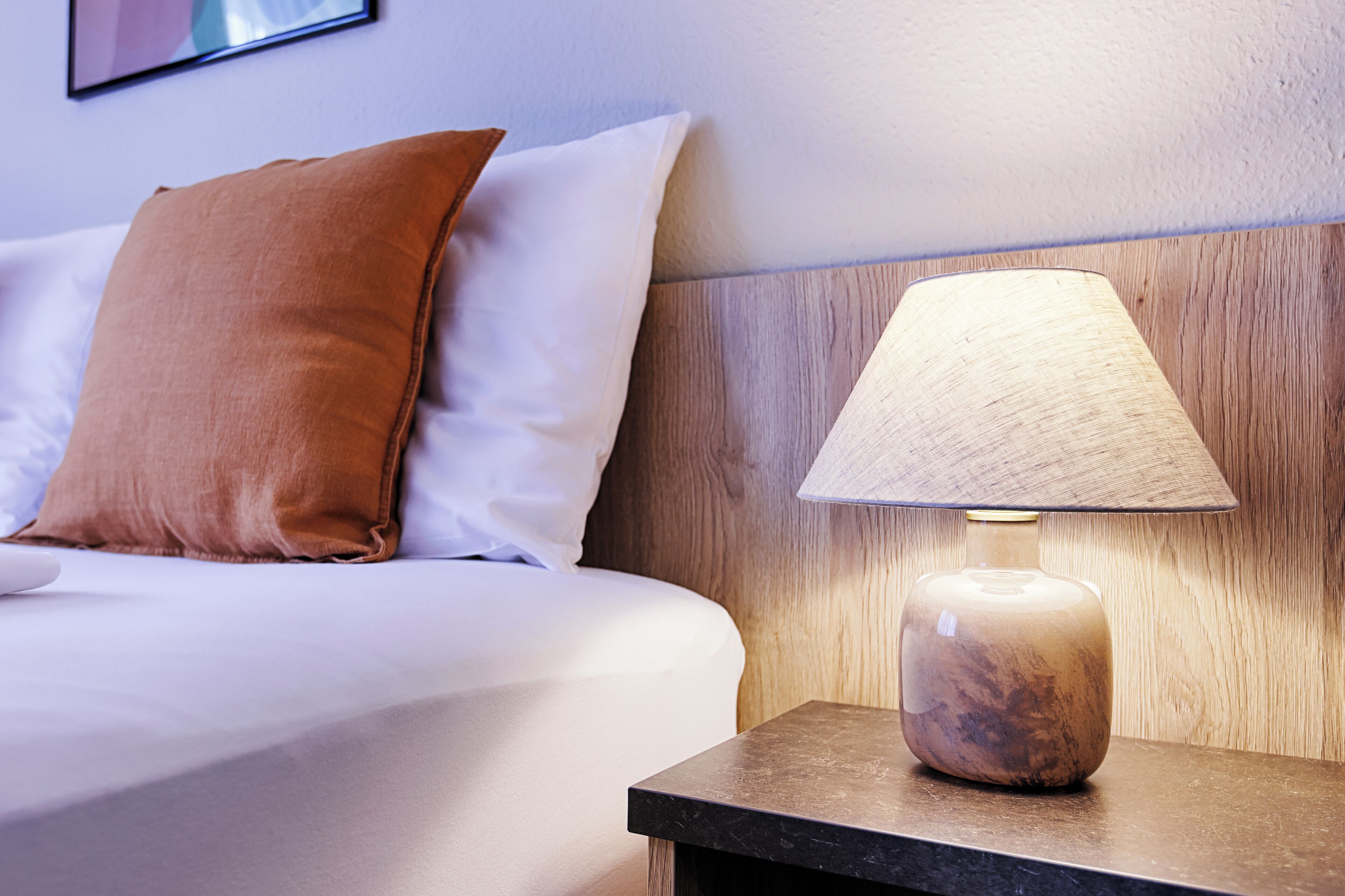 Una cama bien hecha con almohadas blancas y marrones, un cabecero de madera y una mesita de noche con una lámpara.