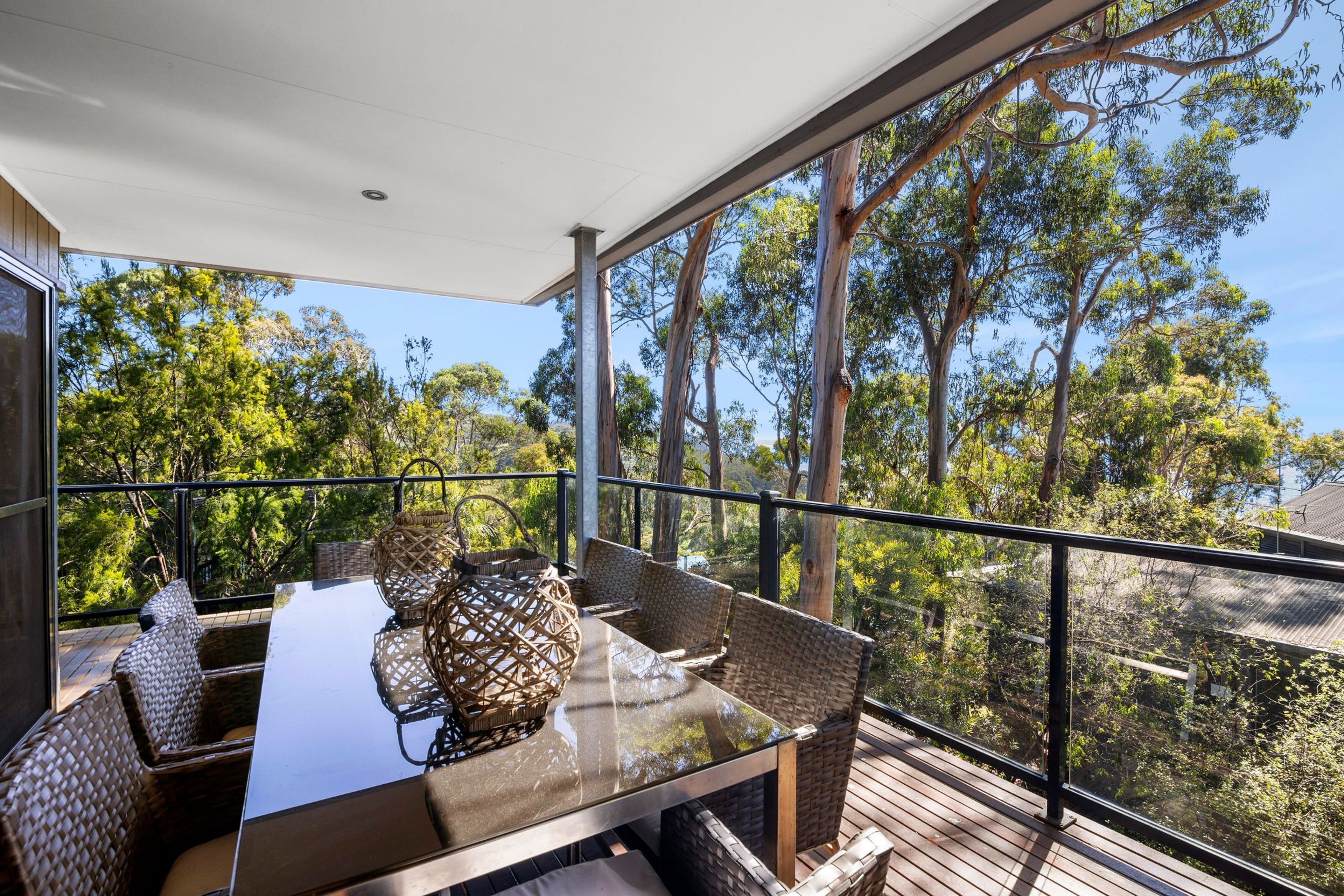 An outdoor balcony with a glass table and wicker chairs, overlooking a forested area.