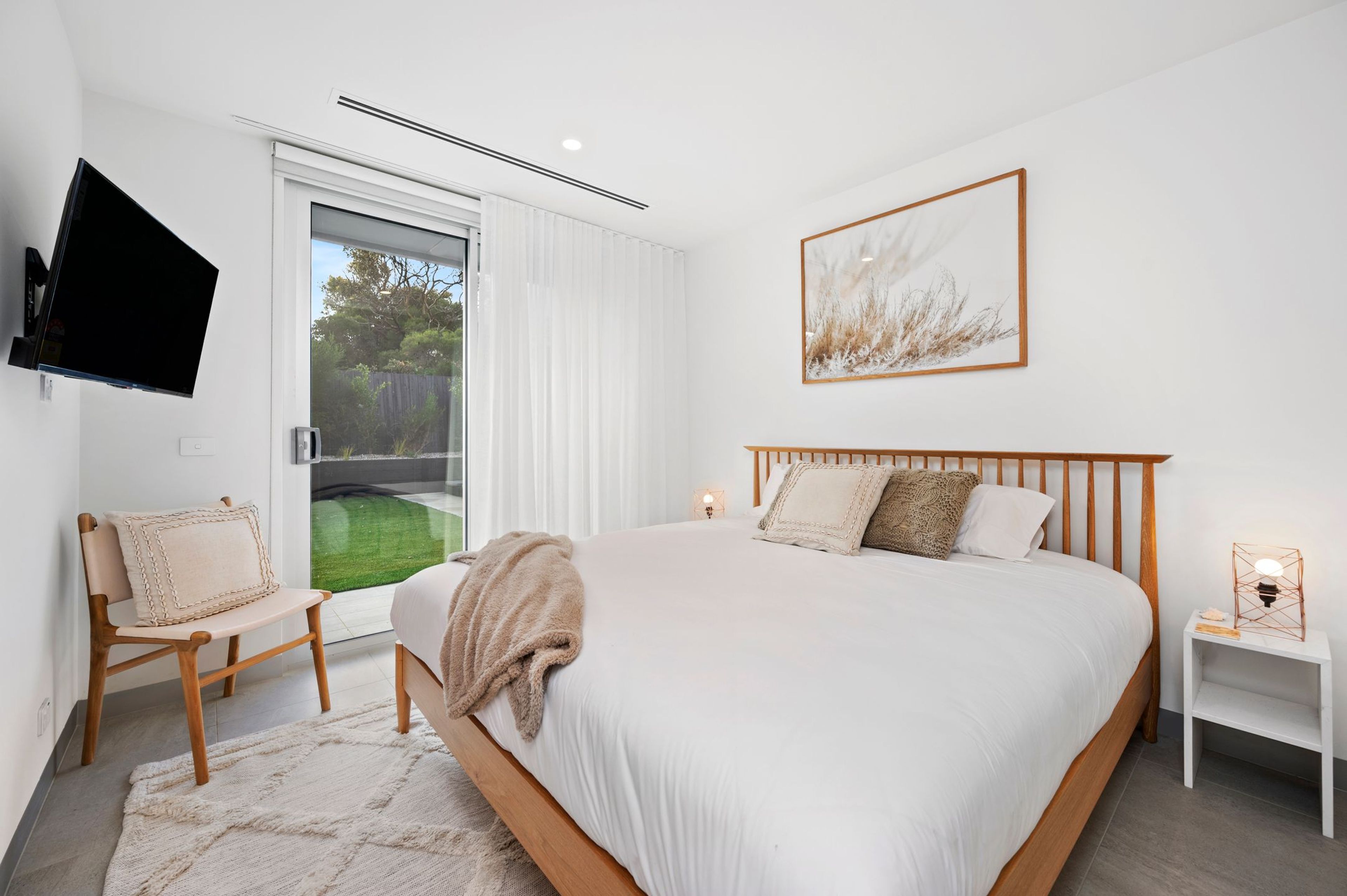 A bedroom with a bed, a chair, a bedside table, a TV mounted on the wall, and a window with curtains.