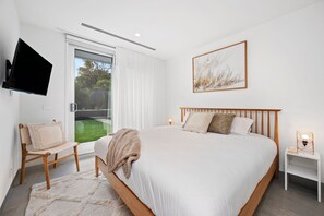 A bedroom with a bed, a chair, a bedside table, a TV mounted on the wall, and a window with curtains.