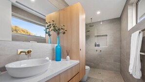 Une salle de bain moderne avec un grand lavabo blanc, un meuble-lavabo en bois, un vase bleu orné de plantes vertes, un espace douche et une vue sur l’extérieur.