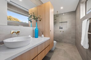 Een moderne badkamer met een groot wit wastafel, een houten wastafelmeubel, een blauwe vaas met groen, een douchegedeelte en uitzicht op het omliggende landschap.