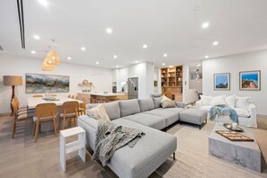 A modern living room with a grey sectional sofa, a dining area with wooden chairs, a kitchen with white cabinets, and a bookshelf.