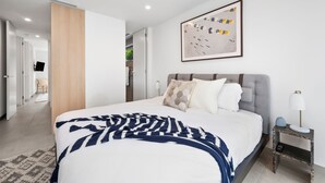 Une chambre à coucher moderne avec un grand lit, une table de chevet, une lampe fixée au mur et une œuvre d’art encadrée.