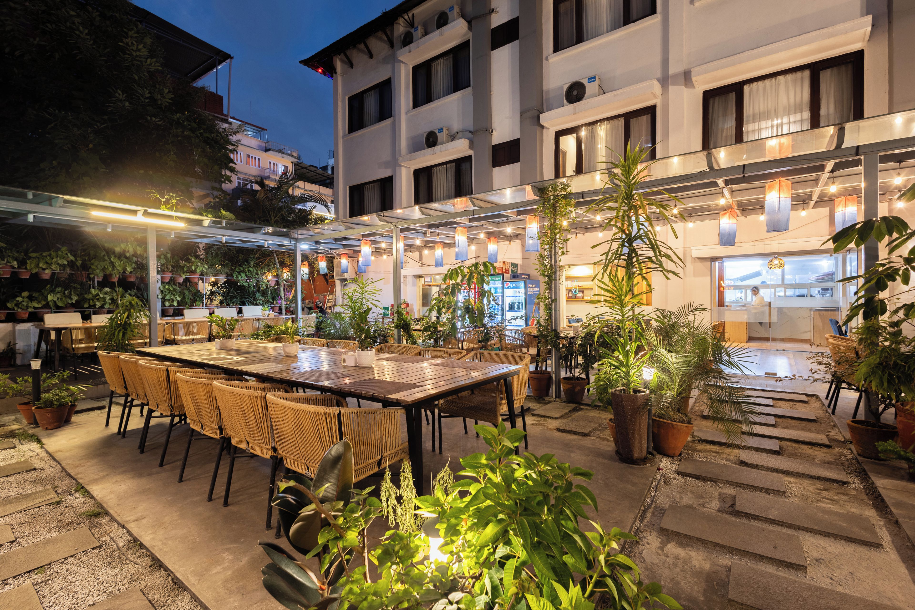 An outdoor dining area with tables and chairs, surrounded by potted plants and a building in the background.