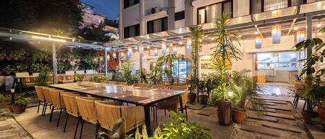 An outdoor dining area with tables and chairs, surrounded by potted plants and a building in the background.