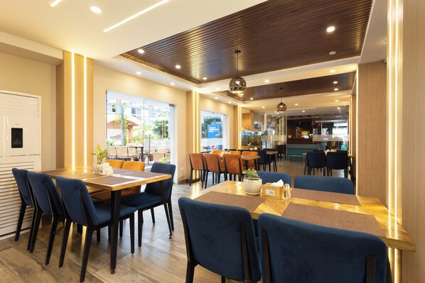 A modern dining area with wooden tables and chairs, a visible kitchen area in the background, and a large window offering a view of the outside.