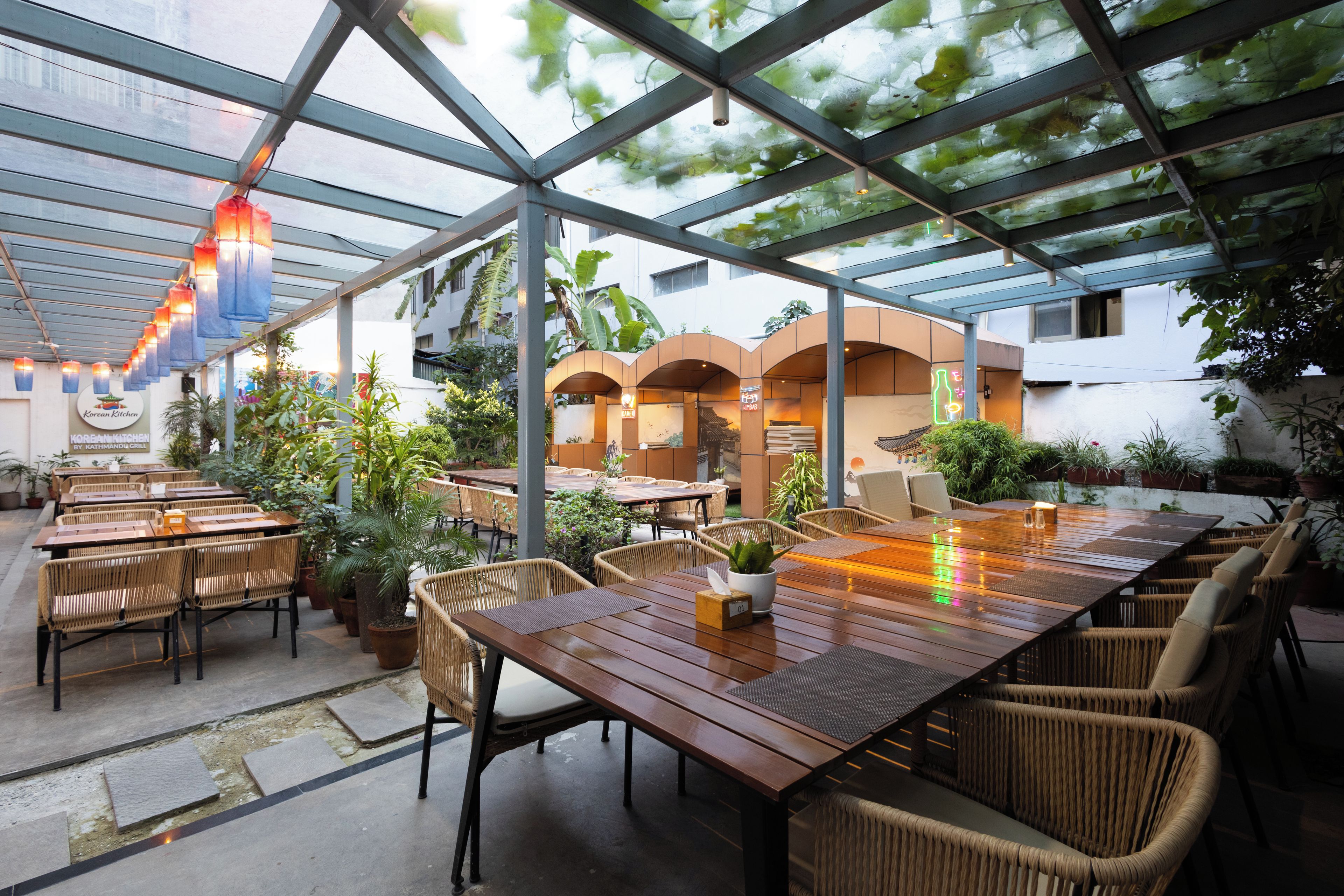 An outdoor dining area with wooden tables and wicker chairs, surrounded by potted plants and a glass ceiling structure.