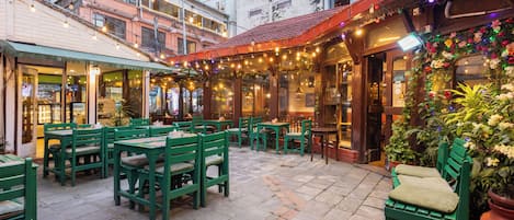 An outdoor seating area with green chairs and tables, surrounded by buildings with visible windows and a brick wall.