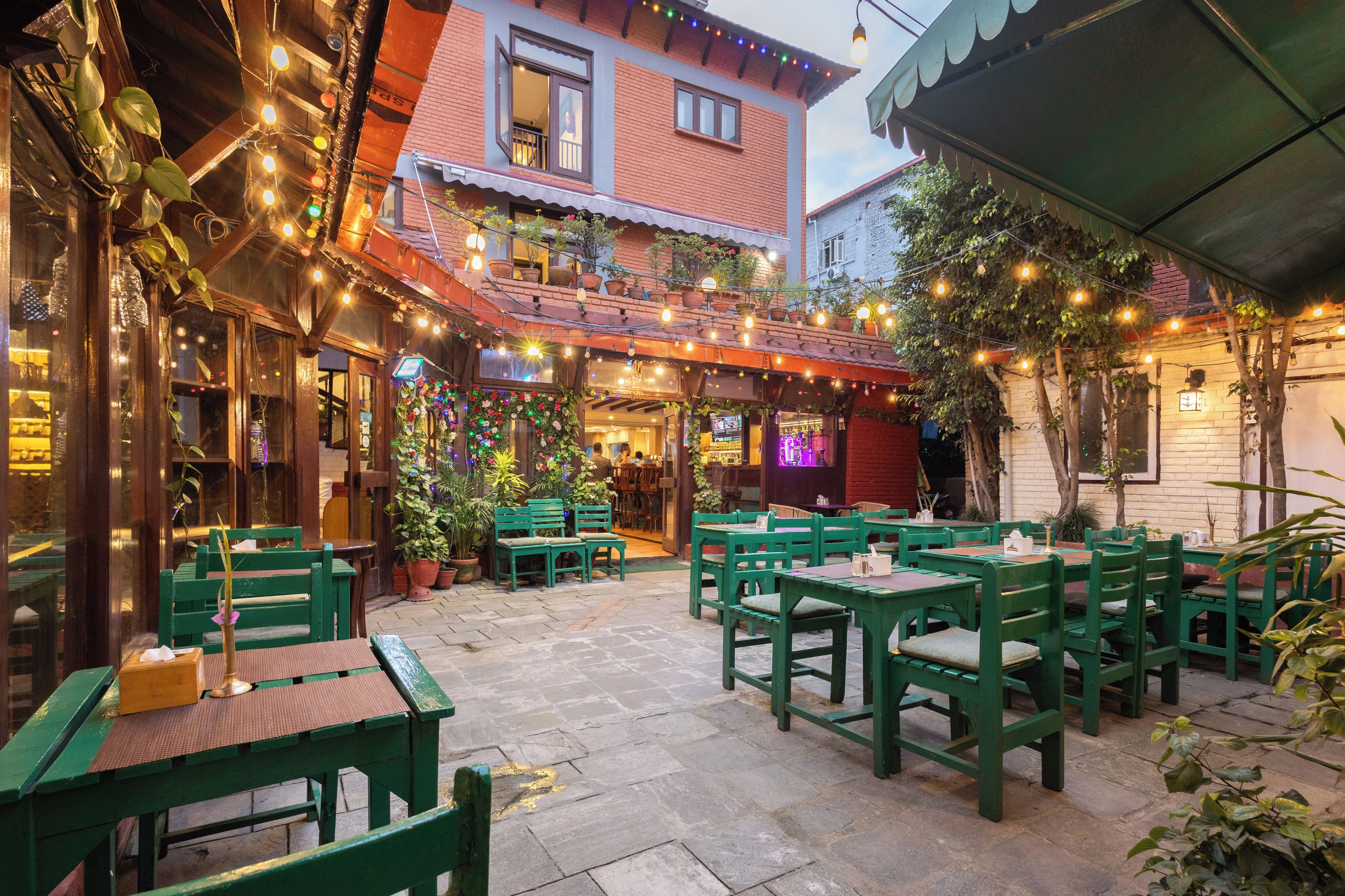 An outdoor seating area with green chairs and tables, surrounded by potted plants and string lights.